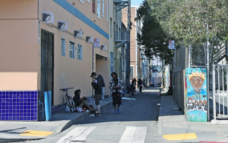 Several people gather on a sidewalk beside a building; graffiti, street signs, and bikes are also visible in the urban alley scene.