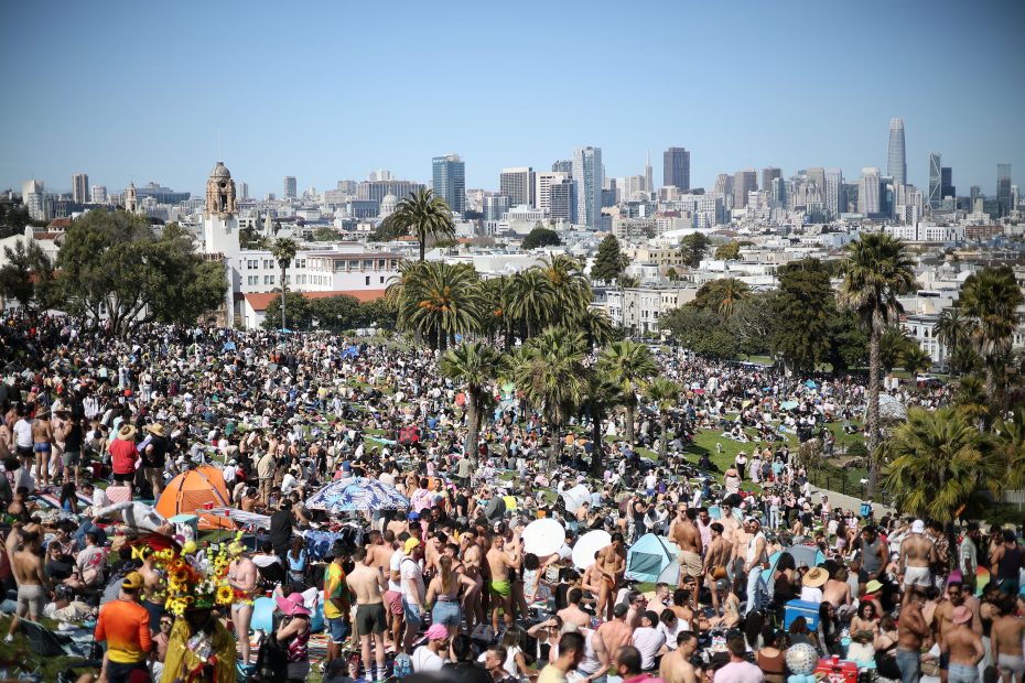 A large crowd gathers in a city park on a sunny day with palm trees and tall buildings visible in the background.
