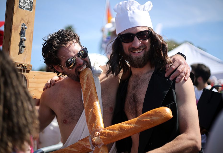 Two men in costumes, one wearing a chef hat and holding a large fake baguette, the other pretending to bite the baguette, posing outdoors at the hunky Jesus competition.
