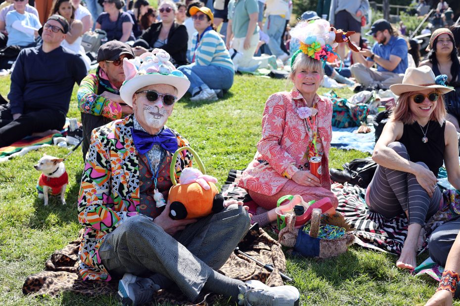 People dressed in colorful, eccentric costumes sit on blankets in a grassy outdoor area, attending an event or festival with others in casual attire around them.