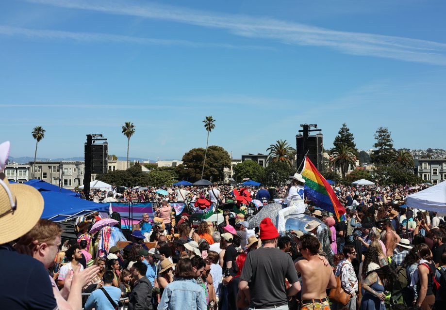 A large crowd gathers outdoors on a sunny day, with tents, palm trees, and a rainbow flag visible among people in colorful clothing.