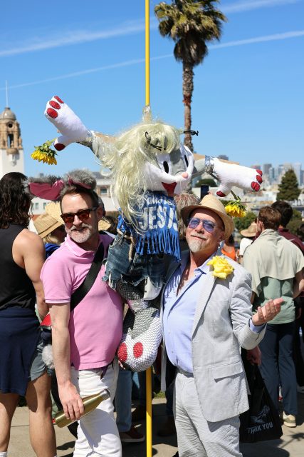Two men pose and smile in front of a colorful, human-sized bunny puppet wearing sunglasses and a "Jesus" shirt at an outdoor gathering, with a crowd and palm tree in the background.