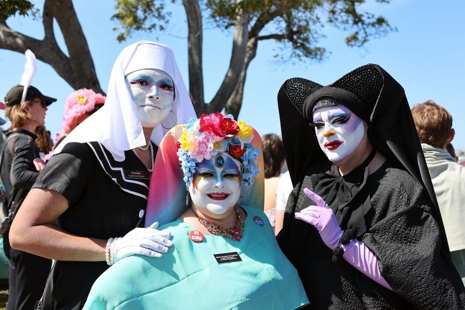 Three people dressed in colorful nun-inspired costumes and white face paint pose together outdoors at a public event.
