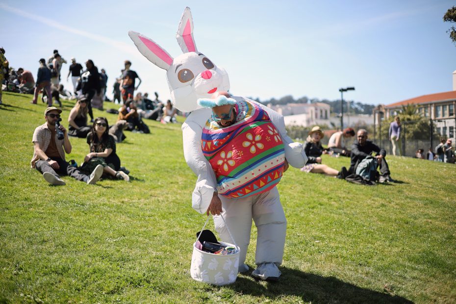 Person in an inflatable Easter Bunny costume holding a decorated basket stands on a grassy hill while people sit and relax in the background on a sunny day.