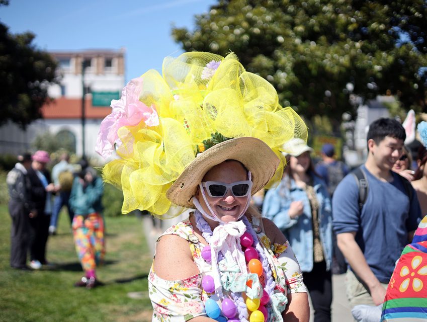 Person outdoors wearing a large yellow hat with decorative tulle, sunglasses, floral shirt, and colorful bead necklace; other people and trees are visible in the background.