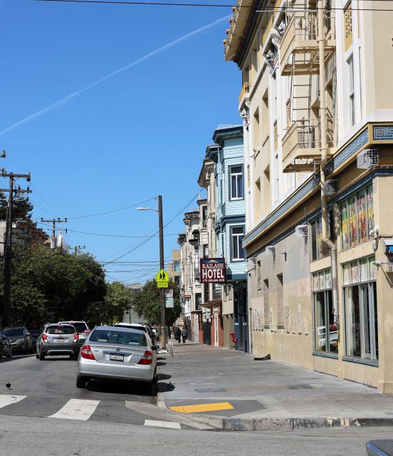 A city street with parked cars, a pedestrian crossing sign, and a hotel sign on a building under a clear blue sky.