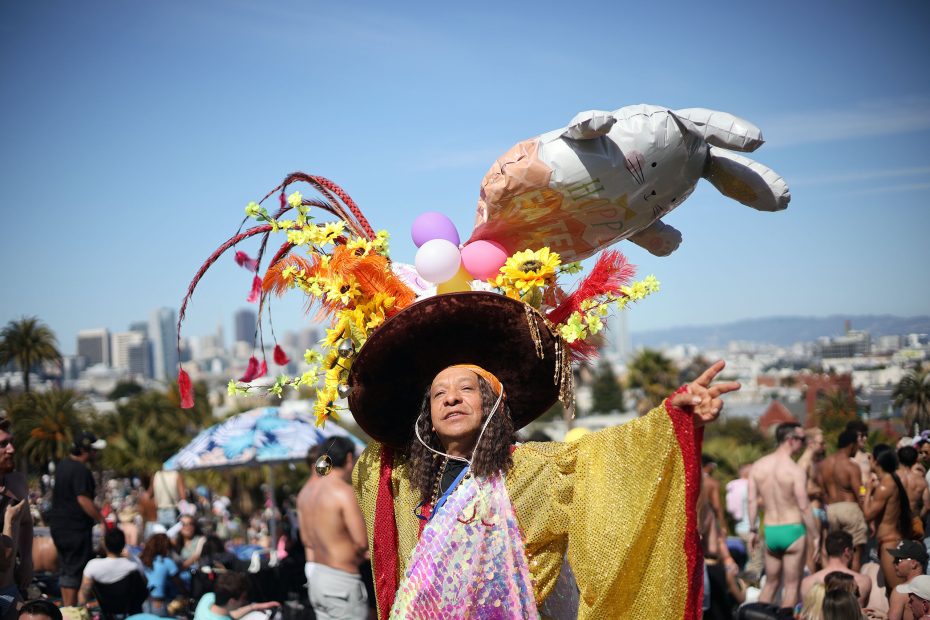 Person in a yellow robe and elaborate hat with flowers, balloons, and an inflatable object poses at an outdoor event in a park with people and city buildings in the background.