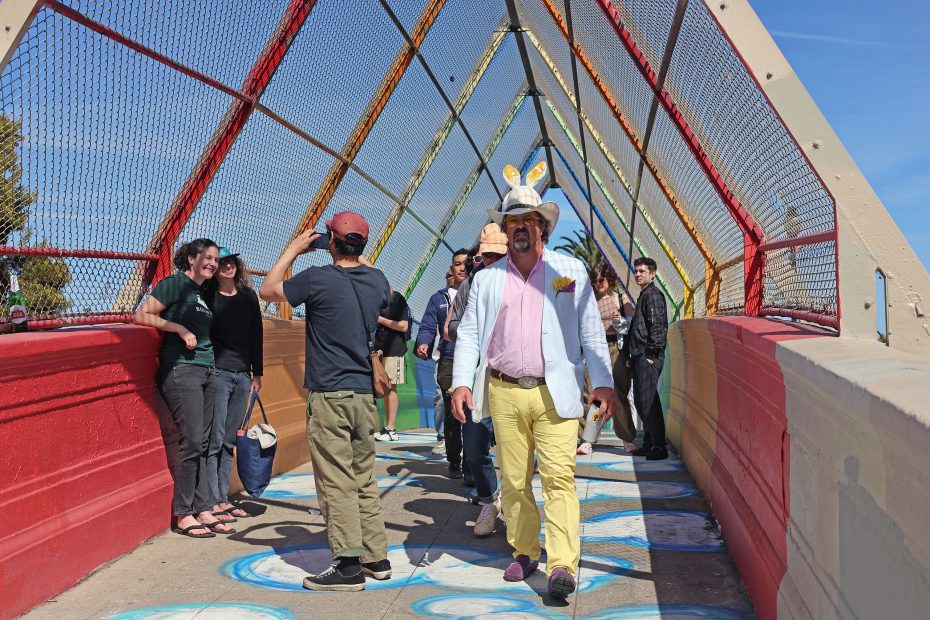 A man in a light blue blazer and yellow pants walks on a colorful pedestrian bridge, surrounded by people, some taking photos, under a bright sky.