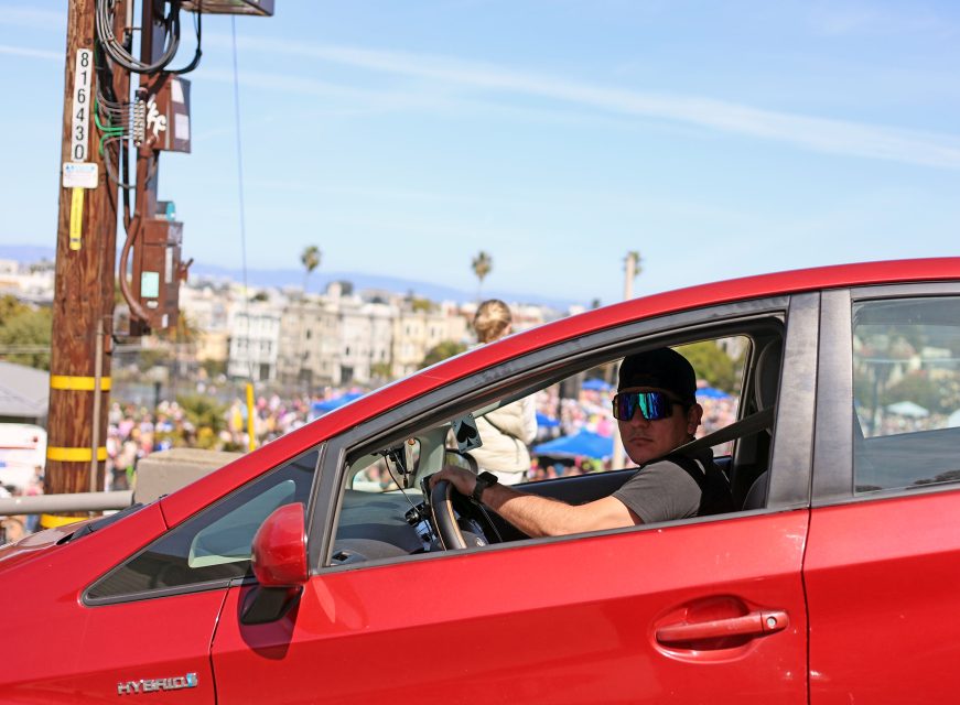 A person wearing sunglasses and a cap drives a red hybrid car with the window down on a sunny day in an urban area.