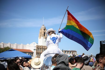 A person in white clothing holds a rainbow pride flag above a crowd during an outdoor event, with a historic building visible in the background.
