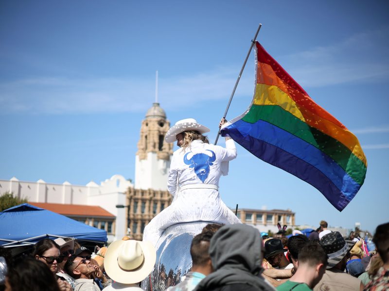 A person in white clothing holds a rainbow pride flag above a crowd during an outdoor event, with a historic building visible in the background.