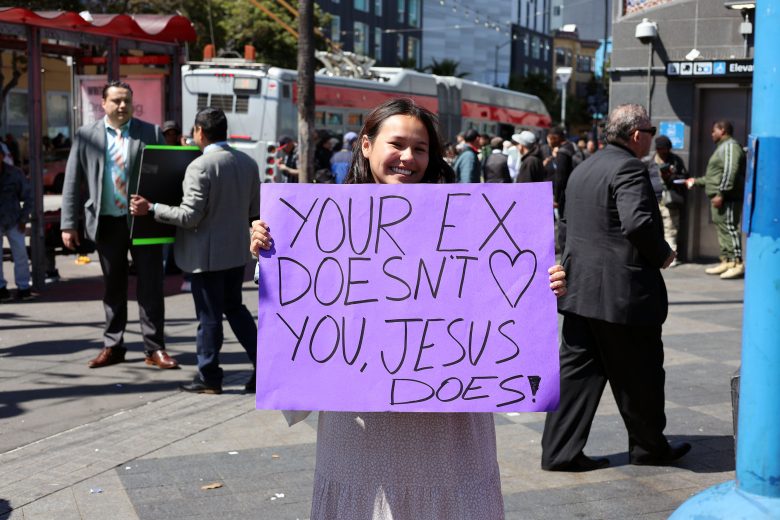 A woman stands in a busy outdoor area holding a purple sign that reads, "YOUR EX DOESN'T LOVE YOU, JESUS DOES!" People walk and talk in the background.