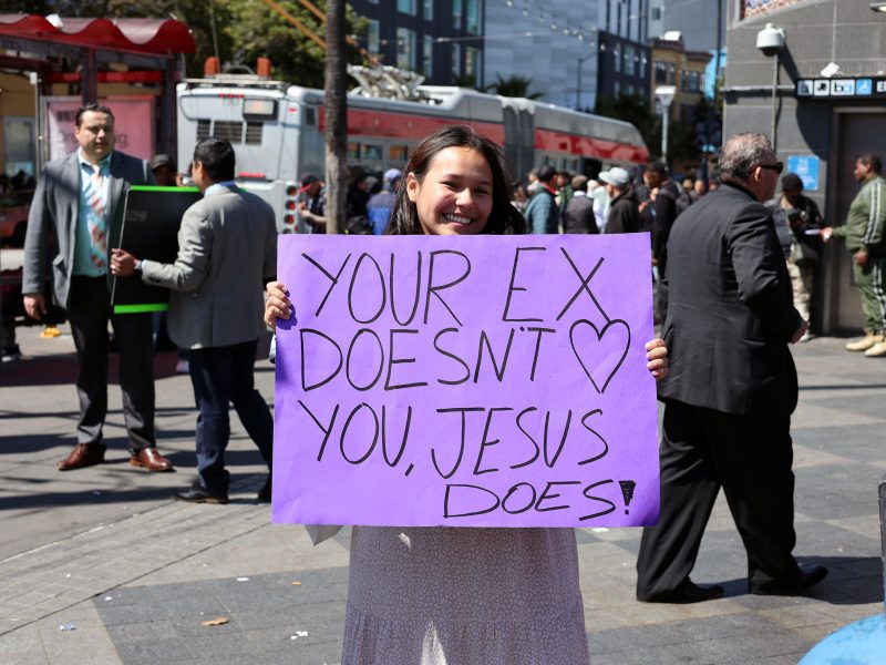 A woman stands in a busy outdoor area holding a purple sign that reads, "YOUR EX DOESN'T LOVE YOU, JESUS DOES!" People walk and talk in the background.