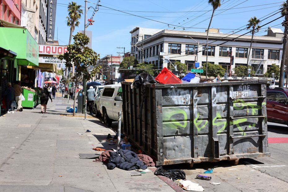 A large dumpster overflowing with trash sits on a city sidewalk next to parked cars, with litter scattered on the ground and storefronts visible in the background.