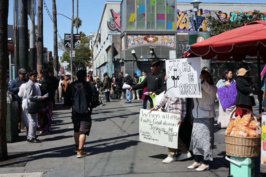 People stand on a busy city sidewalk holding religious signs, with street vendors and colorful murals visible in the background.