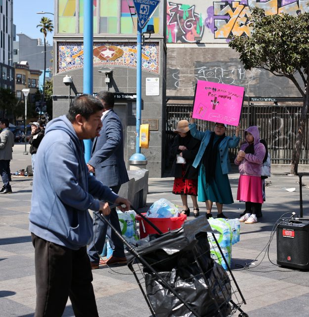 A man pushes a cart in a city plaza while three women stand nearby, one holding a pink protest sign; bags and other items are on the ground.