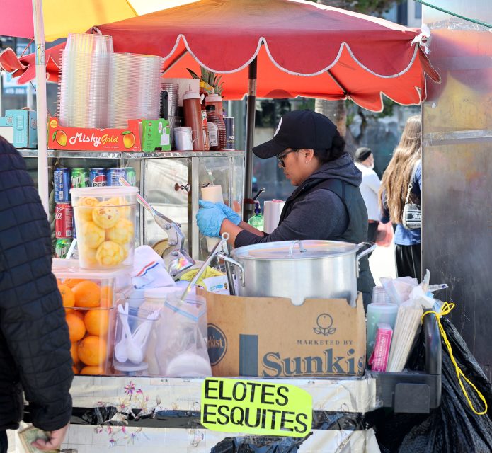 A street vendor wearing a cap and gloves prepares food at a cart stocked with fruits, canned drinks, and a large pot, with signs reading "ELOTES" and "ESQUITES.
