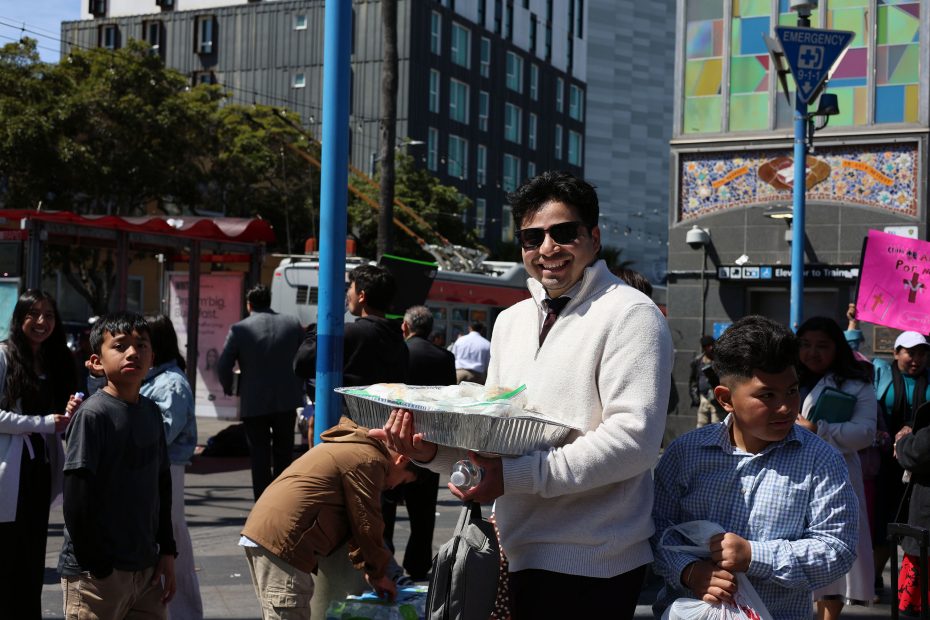 A man in sunglasses holds a large tray of food while standing among a crowd on a city street during the day.
