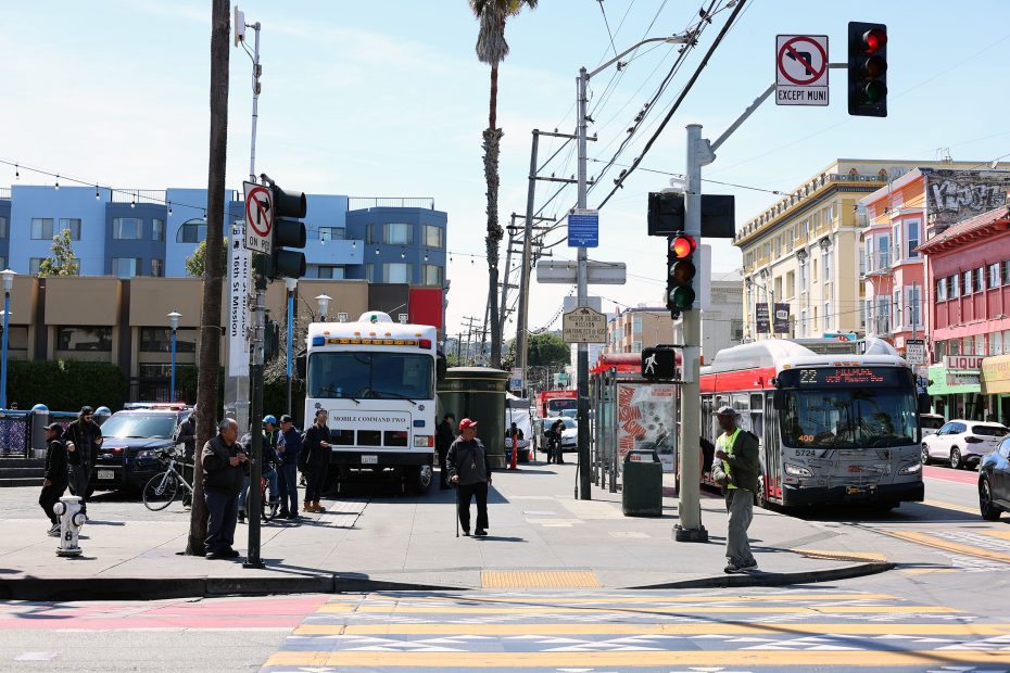 A busy city intersection with pedestrians, a city bus, traffic signals, and various buildings in the background during daytime.