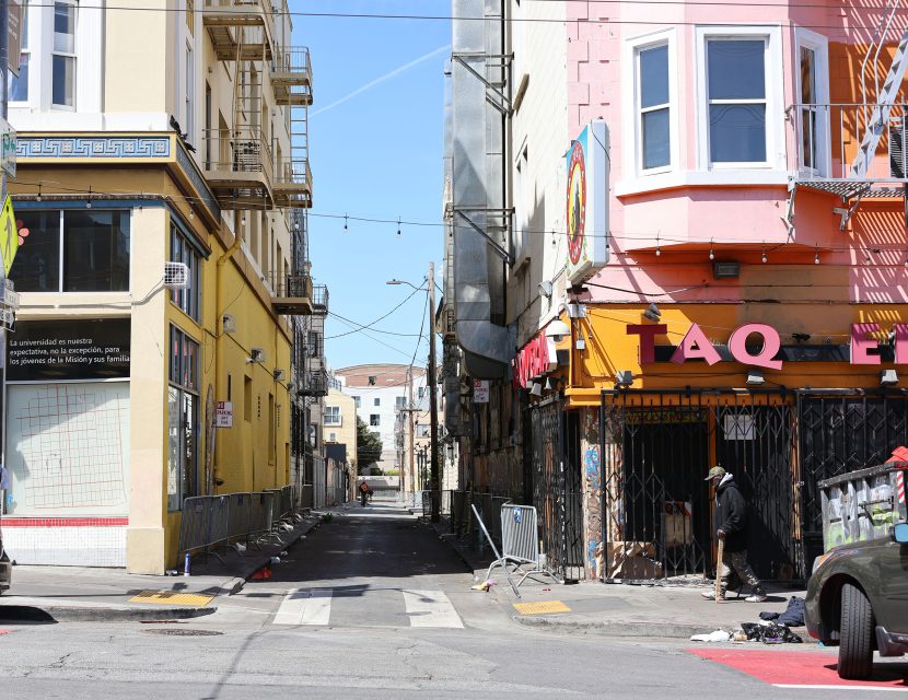 A narrow urban alley between two buildings, with one storefront partially shuttered and debris scattered along the sidewalk. A person stands near the entrance of the alley.