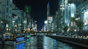 Night view of a canal with illuminated buildings and signs on both sides; boats are visible on the water.