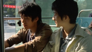 Two young people sit next to each other on a bus, with sunlight coming through the window and empty seats behind them.