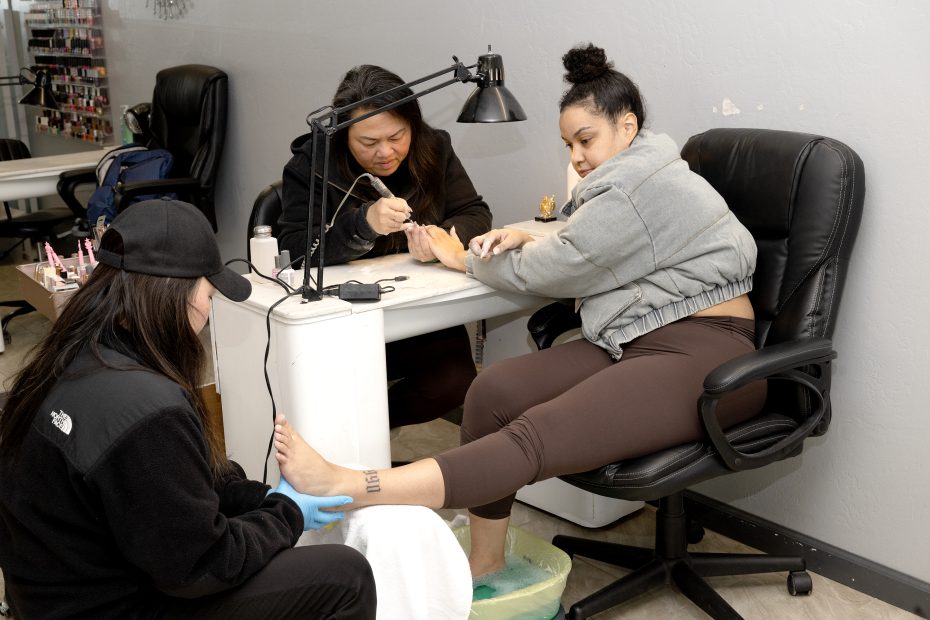Two nail technicians give a pedicure to a woman seated in a black swivel chair inside a nail salon.
