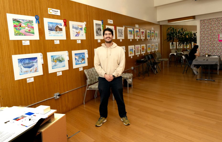 A man stands and smiles in front of a wall displaying colorful artwork and ribbons in a brightly lit room with wood floors and chairs.