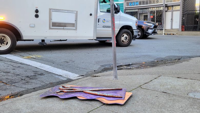 Flattened traffic cones on sidewalk near a pole; white service truck and car visible on the street.