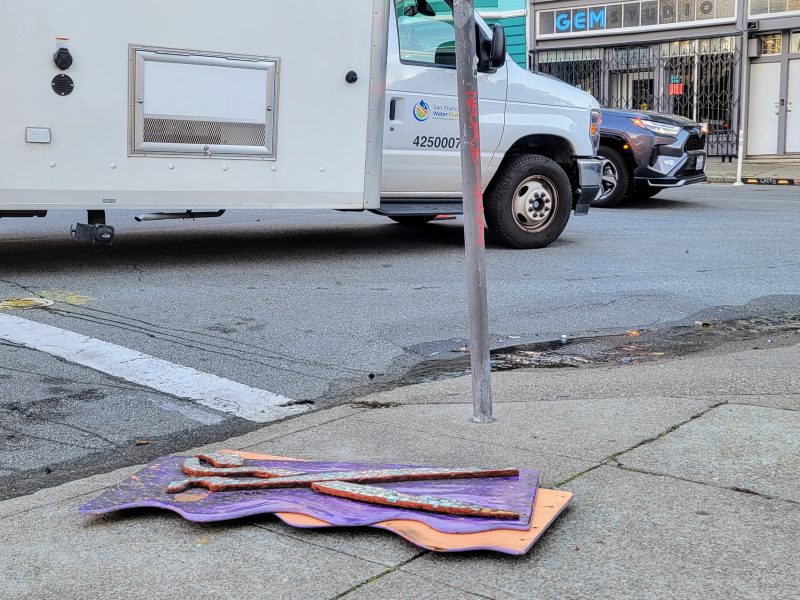 Flattened traffic cones on sidewalk near a pole; white service truck and car visible on the street.