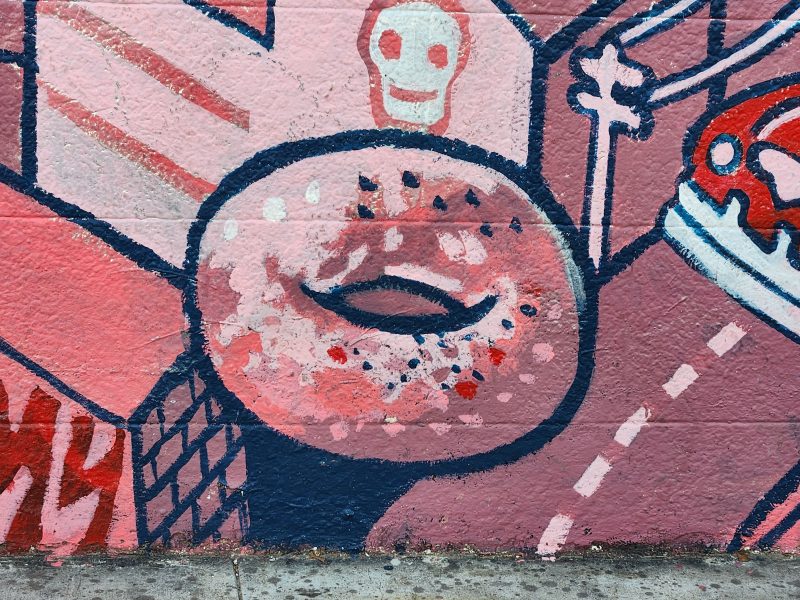 Colorful mural with a central doughnut shape, surrounded by abstract designs, including a skull and various geometric patterns, painted on a brick wall.