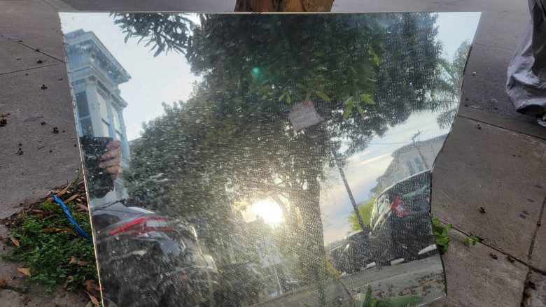 A large rectangular mirror placed on the ground reflects the street scene with cars, trees, and the sun setting in the sky.