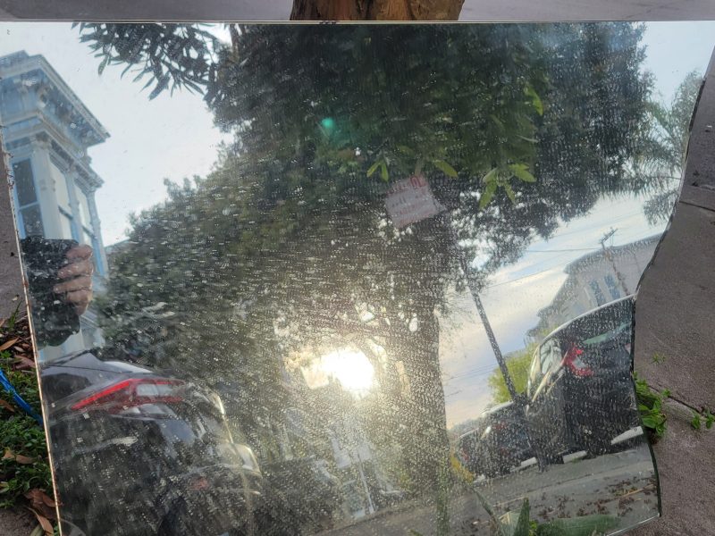 A large rectangular mirror placed on the ground reflects the street scene with cars, trees, and the sun setting in the sky.