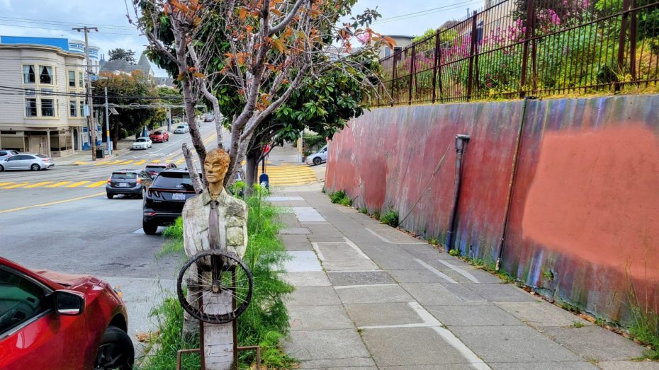 A sidewalk with a weathered mannequin holding a steering wheel in front of a tree, cars parked nearby, and a colorful painted wall to the right.