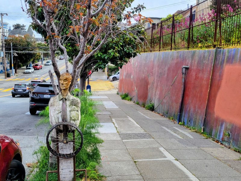 A wooden human figure with a metal steering wheel stands on a sidewalk next to a tree and a colorful wall; cars are parked nearby and a street is visible in the background.