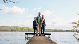 Three men stand on a wooden dock extending over a lake, with green hills and a partly cloudy sky in the background.
