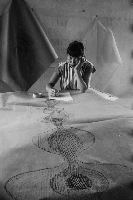 Person sitting at a table, drawing on a large sheet of paper with abstract designs. Other large papers hang in the background.