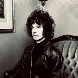 A person with curly hair wearing a dark turtleneck and leather jacket sits on an ornate sofa in a room with framed artwork.