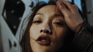 A close-up of a woman’s face, with a hand gripping her head from above. The background is blurry and metallic.