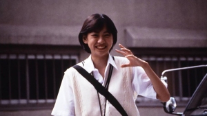 A person with short hair, wearing a white shirt and vest, smiles and waves while standing outdoors near a building and a parked vehicle.