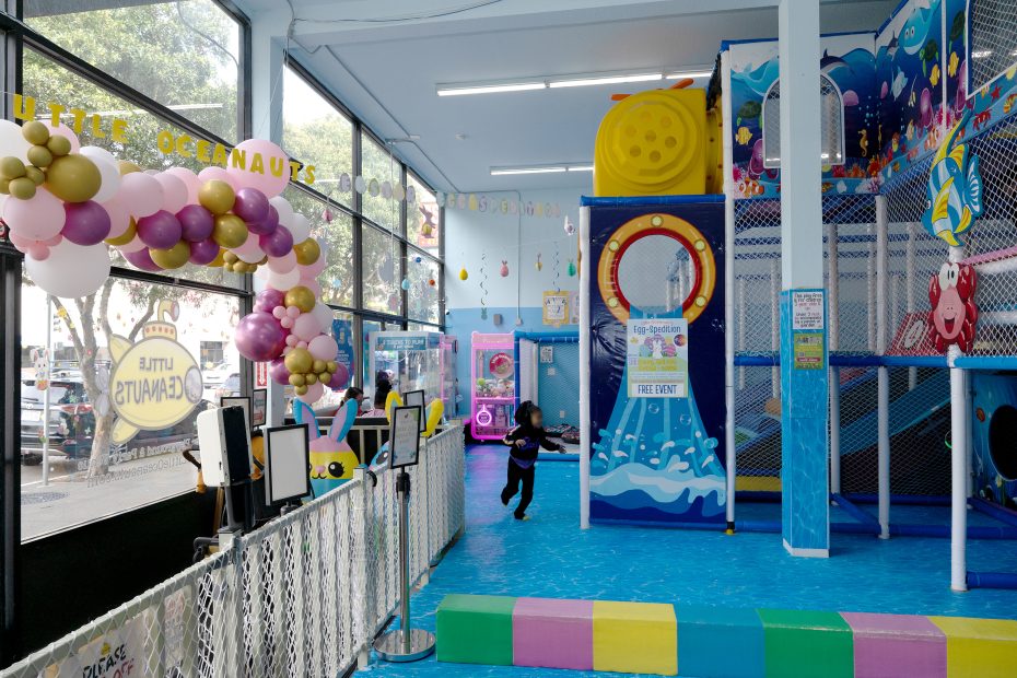 A child runs inside a brightly colored indoor play center with ocean-themed decorations, balloons, and play structures.