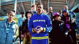 A group of people stands outdoors in front of a stadium, with the central figure wearing a blue "Warrior" tracksuit.