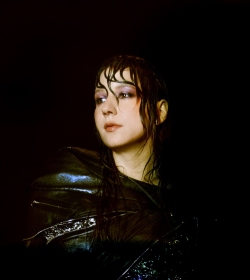 A person with wet hair and dramatic makeup wears a shiny, dark outfit, looking to the side against a dark background.