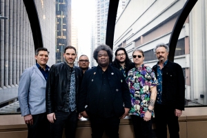 Seven people stand together in a group, wearing a mix of casual and patterned clothing, with a city skyline in the background.