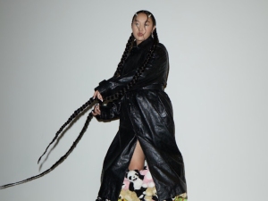 Person wearing a long black leather coat with very long braided hair stands against a plain background; floral-patterned boots are partially visible.