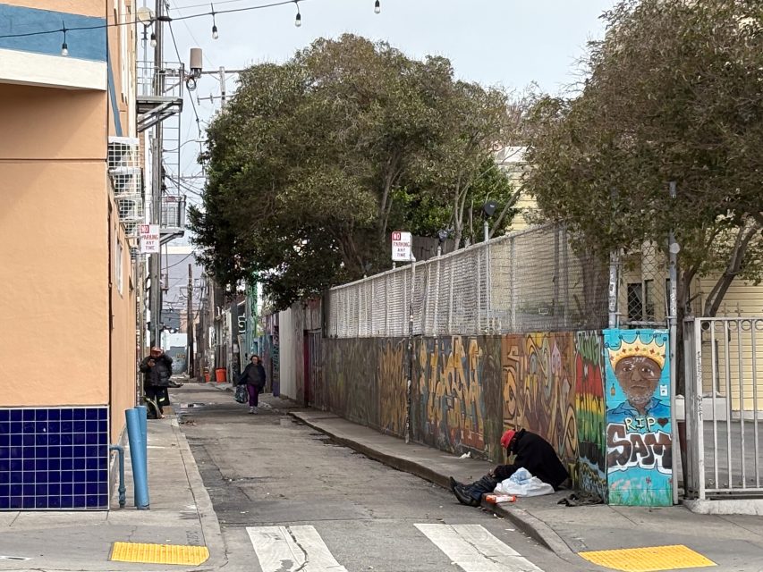 A person sits on the ground in an urban alleyway with graffiti-covered walls, while another person walks further down the alley. Trees and string lights are visible above.
