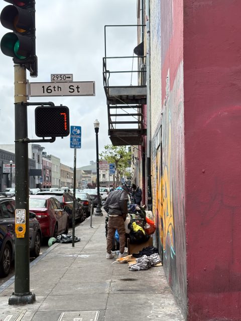A person sorts through belongings on a sidewalk near a building on 16th Street, with cars parked along the street and a walk signal displaying the number 7.