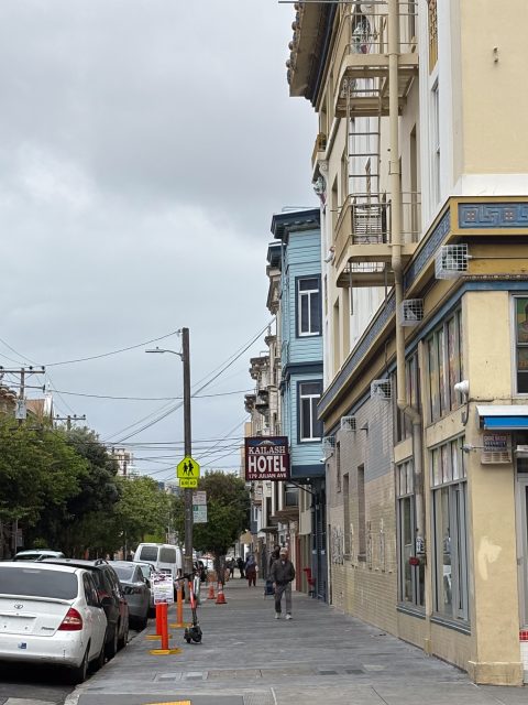 A city sidewalk with parked cars, a person walking, a hotel sign, and buildings under an overcast sky.