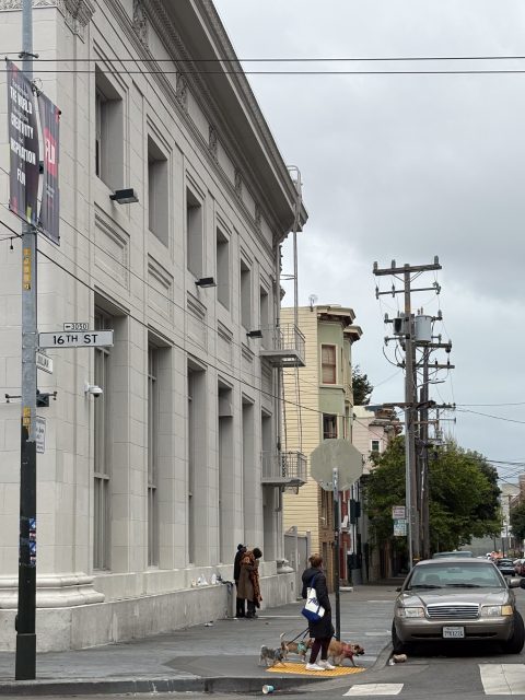 A woman walks three dogs on a leash near the corner of 16th Street, while two people stand talking by the side of a large gray building on a cloudy day.