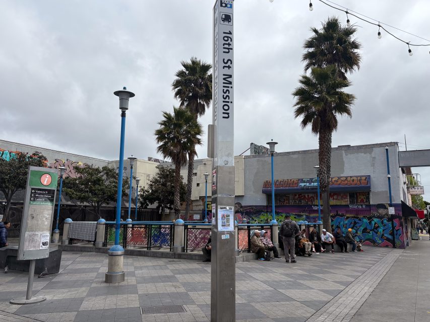 People sit on benches near the 16th St Mission BART station entrance, surrounded by palm trees and buildings with colorful graffiti art.